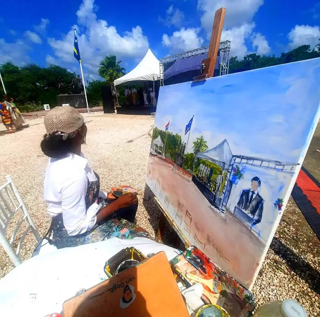 Æmy painting at an easel during an outdoor ceremony, a Curaçao flag beside her