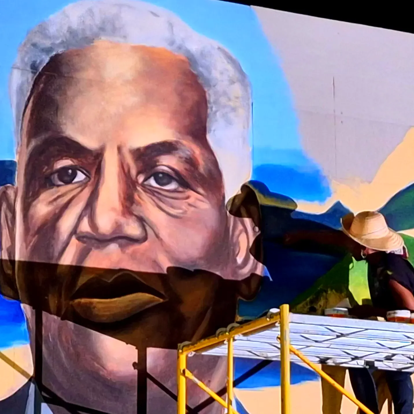 A mural of an old man's face on a building, Æmy at work on the scaffold beside it